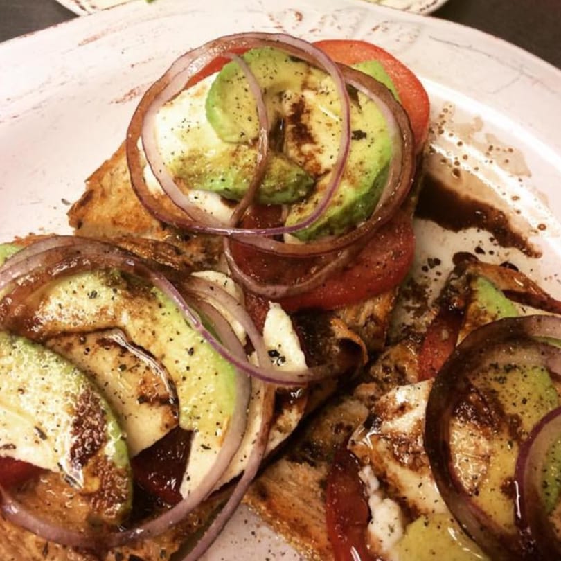 Avocado toast with sliced avocado, red onion, tomato, and balsamic glaze on a white plate.