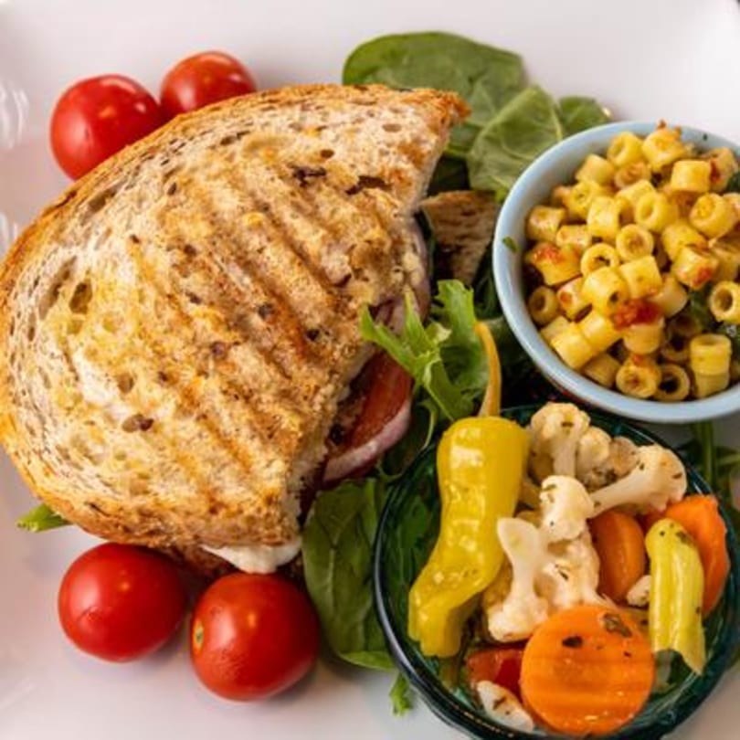 Grilled sandwich with cherry tomatoes, spinach, pasta salad, and pickled vegetables on a white plate.