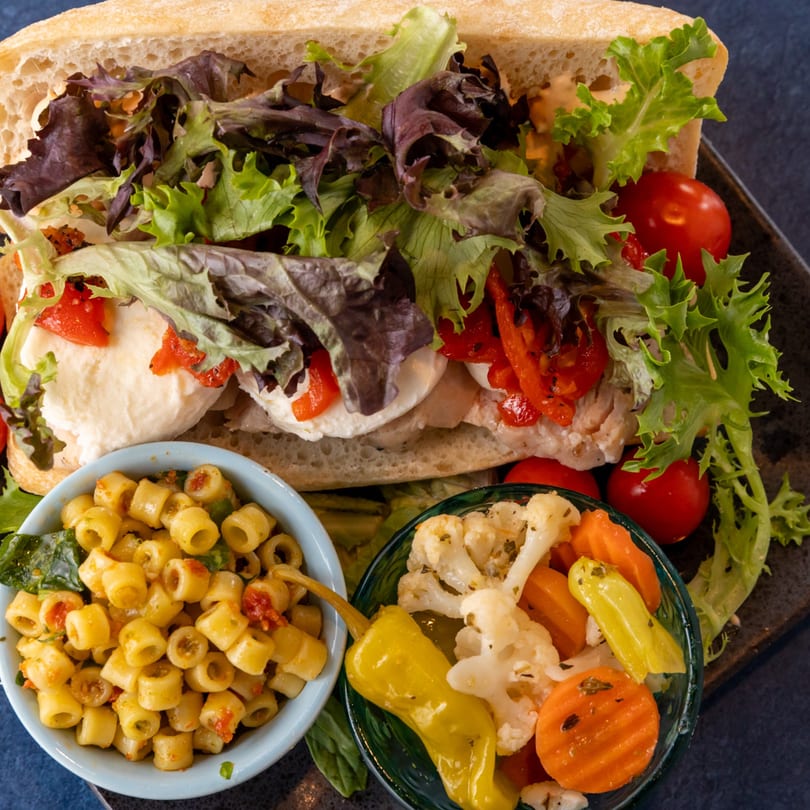 A La Bella Bean sandwich with fresh mozzarella, roasted red peppers, and lettuce, served with pasta salad and pickled vegetables.