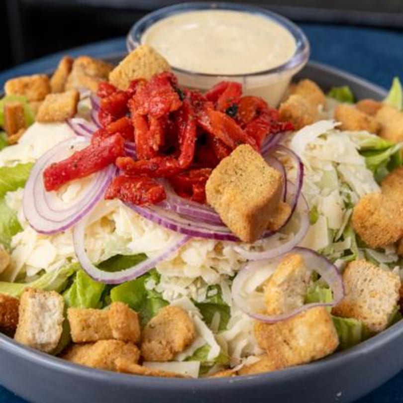 Caesar salad with red peppers, red onions, croutons, and a side of dressing in a bowl.